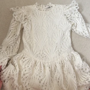 Vit spetsklänning med volang och bälte - Superfin vit klänning i spets från Elsa M. Klänningen har lång ärm, volangdetaljer på axlarna och ett matchande bälte i midjan. Unikt mönster i spetsen och rund halsringning. Perfekt för speciella tillfällen.