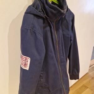 Mörkblå jacka med avtagbar huva, stl för 12-13år - Säljer en mörkblå jacka med huva och hög krage. Jackan har dragkedja framtill och en patch med amerikansk flagga på ena ärmen. Perfekt för vår och höstdagar och har en stilren look.