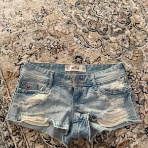 Ljusblå slitna jeansshorts från Hollister - Säljer ett par ljusblå jeansshorts från Hollister med slitna detaljer och råa kanter. Klassisk femficksmodell med låg midja och Hollister-logga på bakfickan. 36 cm i midjemått💕 kommer ha budgivning från 450 kr💕