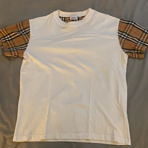 Vit t-shirt med rutiga ärmar från Burberry - Säljer en vit t-shirt från Burberry med klassiska beige, svarta och röda rutiga ärmar. T-shirten har normal passform och korta ärmar. Perfekt för dig som vill ha en stilren look med en twist.