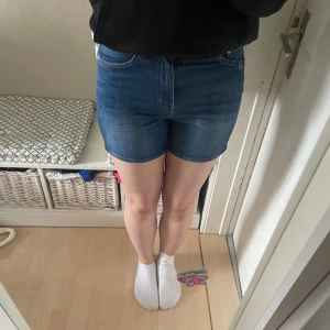 Blå jeansshorts med normal passform - Säljer ett par klassiska blå jeansshorts med fem fickor och knappgylf. Shortsen har en normal passform och är tillverkade i denim med lätt tvättad look. Perfekta för sommardagar och enkla att matcha med olika toppar.