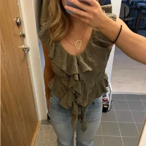 Olivgrön volangtopp från Zara - Säljer en olivgrön topp från Zara med volangdetaljer framtill och djup v-ringning. Toppen är ärmlös och har en strukturerad, lätt skrynklig yta. Perfekt för dig som gillar unika detaljer och en feminin stil.