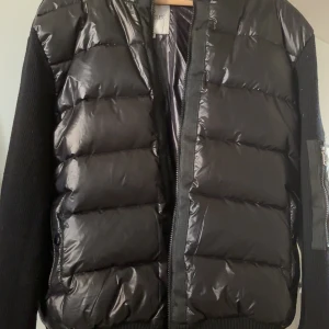 Svart pufferjacka från Moncler - Säljer en svart pufferjacka från Moncler med glansig framsida och stickade ärmar. Jackan har dragkedja och Moncler-logga på ärmen. Perfekt för kyliga dagar och riktigt snygg till streetwear.