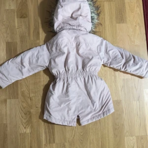 Ljusrosa parkajacka med fuskpäls - Snygg ljusrosa parkajacka med huva och fuskpäls runt kanten. Jackan har dragkedja framtill, knappar och två fickor. Midjan är markerad med resår för en fin passform. Perfekt för kyligare dagar.