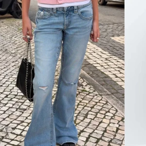 Ljusblå bootcut jeans med slitningar - Säljer ett par ljusblå bootcut jeans med slitningar på knäna. Jeansen har klassisk femficksdesign, normal passform och är tillverkade i denim. Perfekta för dig som gillar en avslappnad och trendig stil. Storlek 164