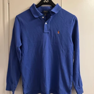 Blå långärmad pikétröja från Polo Ralph Lauren - Säljer en klassisk blå långärmad pikétröja från Polo Ralph Lauren. Tröjan har en broderad logotyp på bröstet och knappar vid kragen. 