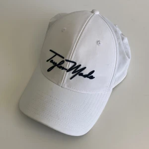 TaylorMade Keps Vit - Snygg vit keps från TaylorMade med svart broderad logga framtill och justerbar rem baktill. Perfekt för golf eller andra utomhusaktiviteter. Klassisk böjd skärm och ventilerande hål för extra komfort.