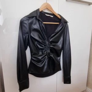 Svart skjorta i skinnimitation från Zara - Säljer en svart långärmad skjorta från Zara i skinnimitation. Skjortan har krage, knappar framtill och snygg rynkdetalj längs knappraden. Perfekt för dig som vill ha en edgy och trendig look.