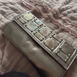 Silvrig kuvertväska med metallplattor - Säljer en snygg silvrig kuvertväska med dekorativa metallplattor på framsidan. Väskan har en avtagbar handledsrem och stängs med dragkedja. Perfekt för att addera lite edge till din outfit!