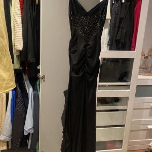 Svart långklänning med paljetter - Elegant svart långklänning med smala axelband och v-ringning. Överdelen är dekorerad med glittrande paljetter och klänningen har en figurnära passform med draperade detaljer längs sidorna. Perfekt för festliga tillfällen.