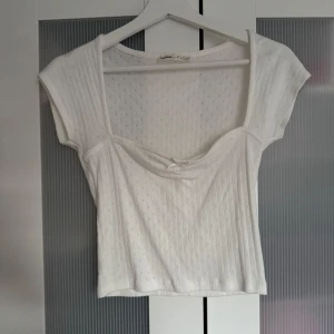 Vit topp med liten rosett - Söt vit topp med små hålmönster och en liten rosett framtill. Toppen har korta ärmar och en djup, fyrkantig urringning. Perfekt för varma dagar. Endast använda en gång. Skriv privat för fler bilder 🩷