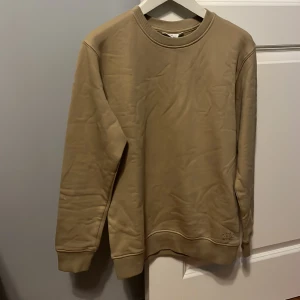 Beige sweatshirt med rund hals - Säljer en stilren beige sweatshirt med rund hals och ribbade muddar. Tröjan har en enkel design utan tryck och passar perfekt till vardags. Mjuk och bekväm med normal passform.