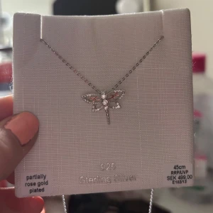 Halsband med trollslända i silver - Säljer ett fint halsband i sterling silver 925 från ur&penn med ett trollslände-hänge dekorerat med gnistrande stenar. Kedjan är 45 cm lång och delvis roséguldspläterad. Man får med original påsen. Fick i konfirmationspresent, tyvärr för sent att lämna tillbaka nu så därför säljer jag🥰. Ordinarie pris 499kr säljer för 470kr aldrig använd! Priset är diskuterbart💘