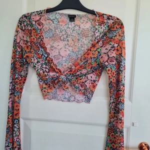 Blommig mesh-top med knyt från Monki - Superfin blommig mesh-top från Monki med långa ärmar och knyt framtill. Toppens färger går i rosa, orange, grön och svart. Perfekt för att addera färg till din outfit!