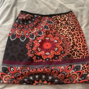 Mönstrad kjol från 101 IDEES - Färgstark kjol med mix av leopardmönster och mandalainspirerade detaljer i rött, svart, orange och grått. Kjolen har en synlig dragkedja bak och är i kort modell. Perfekt för dig som gillar unika prints och vill sticka ut.