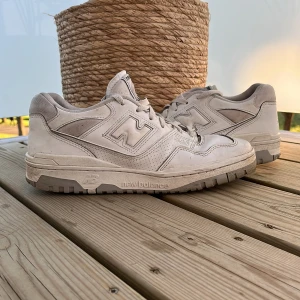 Vita New Balance 550 sneakers - Säljer ett par klassiska New Balance 550 sneakers i vitt med grå detaljer. Skorna har en chunky sula, snörning och den ikoniska N-loggan på sidan. Perfekta för dig som gillar streetwear och sportig stil.