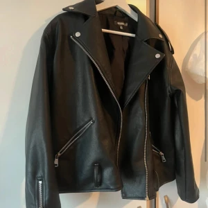 Svart skinnjacka från NA-KD - Säljer en svart skinnjacka från NA-KD med klassisk bikerstil. Jackan har silvriga dragkedjor, bred krage och detaljerade sömmar. I storlek 36 men den är oversize, knappt använd. Men saknar bandet längst ner 