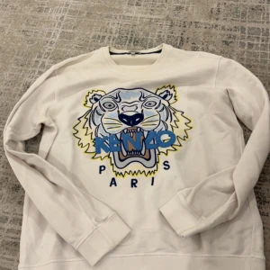 Vit sweatshirt med tigerbrodyr från Kenzo - Säljer en vit sweatshirt från Kenzo med ikonisk tigerbrodyr och blått Kenzo Paris-tryck på bröstet. Tröjan har rund halsringning och långa ärmar. Perfekt statement-plagg för dig som gillar streetwear och snygga detaljer.