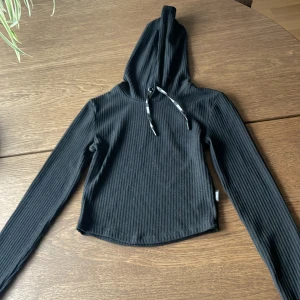 Svart ribbad hoodie från Stronger - Säljer en svart ribbad hoodie från Stronger med långa ärmar och huva. Tröjan har dragsko med logga och är croppad i modellen. Perfekt för en sportig och avslappnad stil.