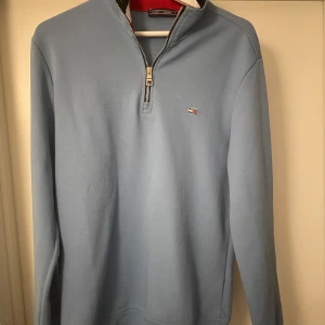 Ljusblå långärmad tröja med dragkedj  från Tommy Hilfiger - Säljer en ljusblå långärmad tröja från Tommy Hilfiger i storlek L. Tröjan har halv dragkedja vid halsen och en liten broderad logga på bröstet. Perfekt för en stilren och avslappnad look.