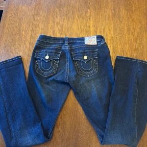 True Religion Joey Low Rise Flare Jeans - Mörkblå jeans från True Religion, modell Joey Low Rise Flare. Byxorna har utställda ben, klassiska kontrastsömmar och dekorativa fickor bak med knappar. Snygg och tidlös modell med låg midja. De är i storlek 28.