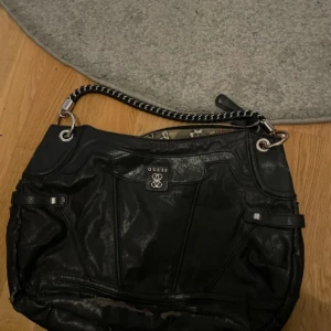 Svart axelväska från Guess med kedjedetalj - Säljer en svart axelväska från Guess med snygg kedjerem i silver. Väskan har flera fack och ett coolt innerfoder med leopard- och blommönster.Men den är ganska sliten bak på väskan och släpper lite från väskan, den går att använda men sliten snygg inuti och frampå!