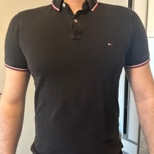 Svart pikétröja från Tommy Hilfiger - Säljer en svart pikétröja från Tommy Hilfiger med klassisk krage och knappar framtill. Tröjan har diskret logga på bröstet och rödvit rand på krage och ärmslut. Storlek L