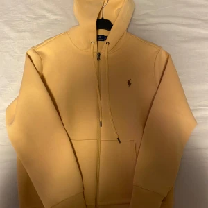 Beige hoodie från Polo Ralph Lauren - Snygg beige hoodie från Polo Ralph Lauren med dragkedja och klassisk logga broderad på bröstet. Tröjan har huva med snörning och två fickor framtill. Perfekt för en avslappnad stil.