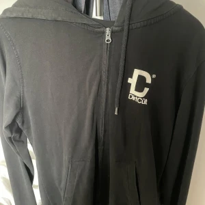 Svart hoodie från DirtCult - Svart hoodie med dragkedja från DirtCult, San Diego. Jackan har huva med dragsko och tryckt logga både fram och bak. Perfekt för en avslappnad stil.