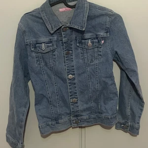 Klassisk blå jeansjacka från Fly - Säljer en klassisk blå jeansjacka från Fly i storlek 146/152. Jackan har knappar framtill, två bröstfickor med lock och knapp samt klassisk krage. Perfekt för dig som gillar tidlös stil och denim.