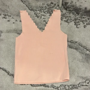 Ljusrosa ribbad linnetopp med sicksackkant - Säljer en ljusrosa ribbad linnetopp med djup v-ringning och sicksackkant längs axelbanden. Toppen är ärmlös och har en mjuk, stretchig känsla. Perfekt att styla till jeans eller kjol.