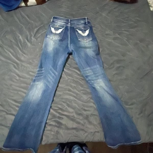Blå bootcut jeans med broderade fickor - Säljer ett par blå bootcut jeans med slitningar och broderade detaljer på bakfickorna. Jeansen har klassisk femficksdesign och markerade sömmar. Perfekta för dig som gillar en retroinspirerad look. Dom är också högmmidjade 