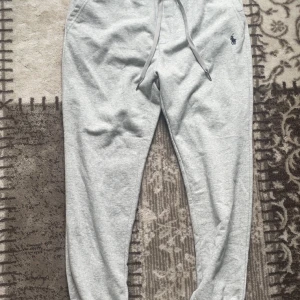 Grå mjukisbyxor från Polo Ralph Lauren - Säljer ett par ljusgrå mjukisbyxor från Polo Ralph Lauren med dragsko i midjan och broderad logga på ena benet. Byxorna har en normal passform och är perfekta för en avslappnad stil.