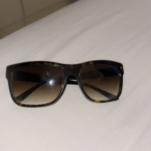 Bruna solglasögon från Versace - Snygga solglasögon från Versace med brunmelerad båge och fyrkantig form. Glasen är tonade i brunt och bågarna har en diskret sköldpaddsmönstrad look. Perfekt accessoar för soliga dagar.