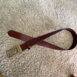 Brunt bälte från donken  - Säljer ett brunt bälte i skinn med ett unikt guldfärgat spänne som har en stilren design. Perfekt accessoar för att lyfta din outfit. Bältet har klassiska hål för justering och passar till många olika stilar.