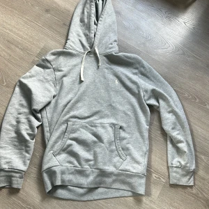 Grå hoodie från Polo Ralph Lauren - Säljer en klassisk grå hoodie från Polo Ralph Lauren med broderad logga på bröstet. Tröjan har känguruficka, dragsko i huvan och ribbade muddar. Perfekt för en avslappnad stil.