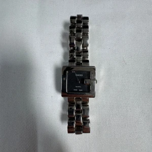 Vintage ’G frame’ Gucci Klocka - Stilren klocka från Gucci med kvadratisk urtavla och länkarmband i silverfärgad metall. Urtavlan är svart med diskreta visare och märkets logga. Klockan har quartz-urverk och är Swiss Made. Perfekt accessoar för dig som gillar klassisk design. (Officiella Gucci lådan är bort tappad så ej includerad)
