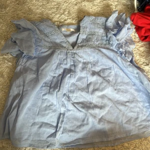 Ljusblå blus med volangärmar från Gina Tricot - Säljer en ljusblå blus från Gina Tricot med v-ringning och dekorativa spetsdetaljer upptill. Blusen har korta volangärmar och är tillverkad i ett lätt bomullstyg. Perfekt för vår och sommar!