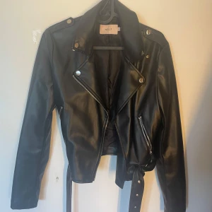 Svart skinnjacka från Nelly - Säljer en svart skinnjacka från Nelly i storlek 34. Jackan har klassisk bikerstil med dragkedjor, knappar och bälte i midjan. Perfekt för dig som vill ha en tidlös och snygg look med coola detaljer.