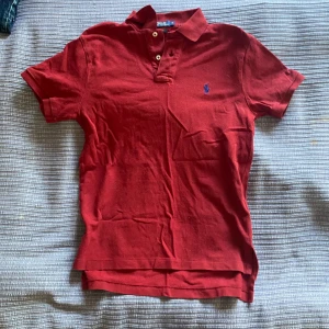 Röd pikétröja från Polo Ralph Lauren - Säljer en klassisk röd pikétröja från Polo Ralph Lauren med liten broderad logga på bröstet. Tröjan har korta ärmar, krage och knappar vid halsen. 