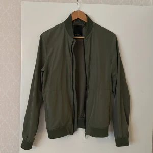 Olivgrön bomberjacka från J.Lindeberg - Snygg olivgrön bomberjacka från J.Lindeberg med dragkedja framtill och ribbade muddar vid ärmslut, krage och nederkant. Jackan har en klassisk, stilren design och passar perfekt till vardagslooken. Perfekt sommarjacka. I mycket bra skick! 