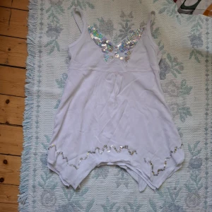 Vit linne-topp med paljettdetaljer - Säljer en vit linne-topp med smala axelband och glittriga paljetter i silver över bysten och längs nederkanten. Toppen har en asymmetrisk nederdel och är perfekt för dig som gillar att sticka ut.