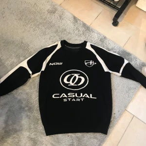 Svart och vit tröja med tryck från Casual Start - Säljer en svart tröja med vita detaljer och tryck från Casual Start. Tröjan har rund halsringning och långa ärmar med sportig design och grafiska loggor på bröstet och ärmarna.