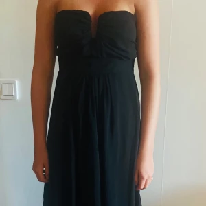 Svart långklänning från Glamorous S - Säljer en svart axelbandslös långklänning från Glamorous. Klänningen har en draperad byst och markerad midja, vilket ger en elegant siluett. Perfekt för speciella tillfällen när du vill känna dig extra fin.