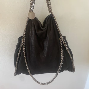 Svart axelväska från Stella McCartney - Säljer en snygg svart axelväska från Stella McCartney med kedjedetaljer i silver. Använd få tal gånger och kom med lappar när jag köpte den. Kan säljas men vill helst byta mot en liten mindre storlek 