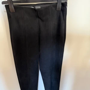 Svarta byxor i mockaimitation från Zara - Snygga svarta byxor från Zara i mjuk mockaimitation. De har hög midja, smal passform och dragkedja i sidan. Perfekta till en stilren look och enkla att matcha med olika toppar.