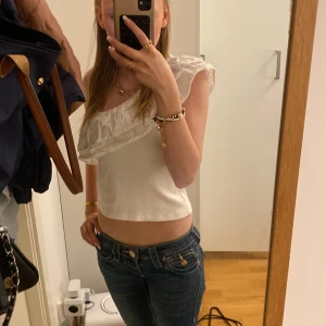 Vit omlott topp med volang från Gina Tricot - Säljer en söt vit omlott topp från Gina Tricot med volangdetalj över axeln. Toppen är ärmlös och har broderade spetsdetaljer som ger en romantisk känsla. Perfekt till sommaren!