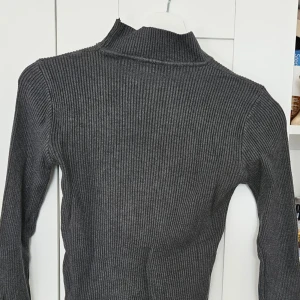 Mörkgrå ribbad polotröja - Säljer en mörkgrå ribbad polotröja med lång ärm. Tröjan är figurnära och har en klassisk polokrage, perfekt för lager-på-lager under kyligare dagar.