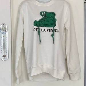 Vit sweatshirt från Bottega Veneta med grön sko-tryck - Säljer en vit sweatshirt från Bottega Veneta med ett stort grönt tryck av en sko på bröstet och gröna snören. Tröjan har rund halsringning och långa ärmar. Perfekt för dig som gillar streetwear och vill sticka ut.