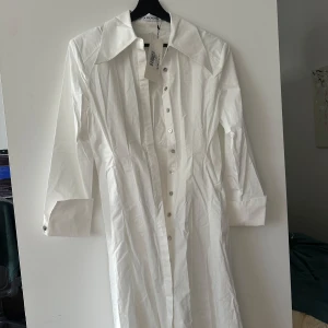 Vit långärmad klänning från ADOORE - Snygg vit långärmad klänning från ADOORE med knappar framtill. Perfekt för en stilren look. Klänningen har en klassisk krage och är tillverkad i ett lätt material.